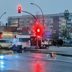 Verkehrsunfall mit mehreren Fahrzeugen an einer Kreuzung, nasse Straße, mehrstöckiges Gebäude im Hintergrund.