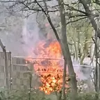 Flammen schlagen hinter einem Zaun auf, umgeben von Bäumen und Rauch.
