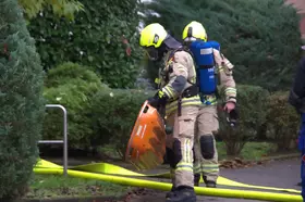 Zwei Feuerwehrleute in Schutzkleidung und gelben Helmen legen die Schleifkorbtrage auf einem Gehweg neben grünen Büschen ab.
