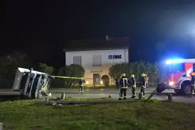 Umgekippter weißer Lkw neben umgestürztem Straßenlaternenmast, Feuerwehrleute in Schutzkleidung und Einsatzfahrzeug mit Blaulicht vor Wohnhaus bei Nacht.