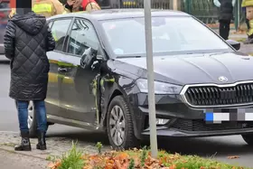 Schwarzes Auto mit beschädigter Seite, daneben steht eine Person in schwarzer Steppjacke und blauen Jeans auf einem Gehweg.