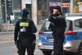 Zwei Polizisten in schwarzer Einsatzkleidung mit gelben "POLIZEI"-Schriftzügen stehen vor einem Polizeiauto in einer Stadtstraße.