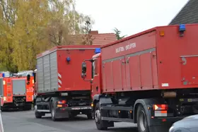 Rote Einsatzfahrzeuge mit blauen Warnleuchten stehen auf einer Straße, im Hintergrund herbstliche Bäume.