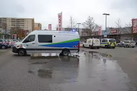 Weißer Transporter der Wasserbetriebe mit blau-grünem Streifen steht auf nassem Parkplatz vor REWE-Markt, ein Einsatzfahrzeug der Polizei im Hintergrund.