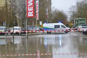 Wasserfläche auf Parkplatz vor REWE-Markt, Absperrband und Einsatzfahrzeug der Wasserbetriebe mit Blaulicht sichtbar.