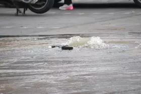 Wasser sprudelt aus einem Kanaldeckel in einer überfluteten Straße, im Hintergrund sind Fahrzeugreifen und ein Fuß mit pink-weißem Schuh sichtbar.