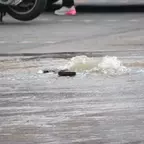 Wasser sprudelt aus einem Kanaldeckel in einer überfluteten Straße, im Hintergrund sind Fahrzeugreifen und ein Fuß mit pink-weißem Schuh sichtbar.