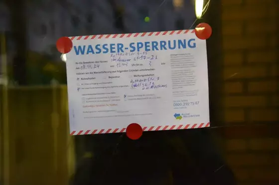 Zettel mit der Überschrift 'WASSER-SPERRUNG' und handschriftlichen Notizen, befestigt mit roten Klebepunkten an einer Glasfläche.