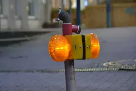 Gelbe Warnleuchte mit zwei orangefarbenen Rundlampen an einem Metallrohr mit Wasserhahn auf gepflastertem Boden.