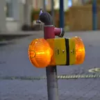 Gelbe Warnleuchte mit zwei orangefarbenen Rundlampen an einem Metallrohr mit Wasserhahn auf gepflastertem Boden.