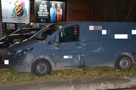 Grauer Transporter steht nachts auf einem Gehweg, sichtbare Schäden an der Fahrertür, im Hintergrund beleuchtete Werbetafeln.