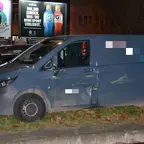 Grauer Transporter steht nachts auf einem Gehweg, sichtbare Schäden an der Fahrertür, im Hintergrund beleuchtete Werbetafeln.