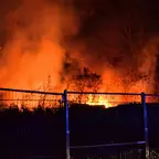 Großes Feuer mit orangefarbenen Flammen und Rauch hinter einem Bauzaun in der Nacht.