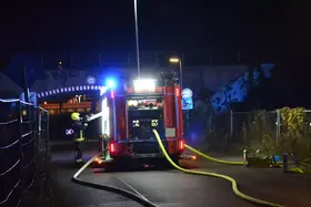 Feuerwehrauto mit eingeschalteten Blaulichtern steht nachts auf Straße, ein Feuerwehrmann bedient sich aus dem Fahrzeug vor einem Tunnel mit Graffiti.