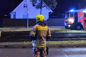 Feuerwehrmann mit gelbem Helm und Schutzkleidung steht vor einem Wohnhaus, im Hintergrund ein Einsatzfahrzeug mit Blaulicht.