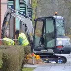 Kleiner grauer Kubota-Bagger mit ausgefahrenem Arm neben zwei Personen in gelben Warnwesten vor einem Wohnhaus.
