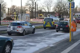 Mehrere Fahrzeuge fahren auf einer Straße mit weißem Bindemittel auf der Fahrbahn, im Hintergrund Baustellenabsperrungen und Bäume ohne Laub.