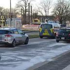 Mehrere Fahrzeuge fahren auf einer Straße mit weißem Bindemittel auf der Fahrbahn, im Hintergrund Baustellenabsperrungen und Bäume ohne Laub.