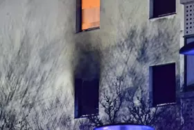 Fassade eines Gebäudes mit mehreren Fenstern, aus einem davon kommt Rauch, Schatten von Ästen auf der Wand sichtbar.