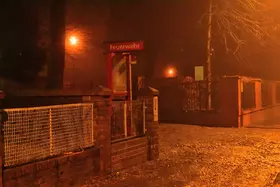 Einfahrt zur Feuerwache bei Nacht mit rotem Schild 'Feuerwehr', Laterne und kahlen Bäumen im Hintergrund.