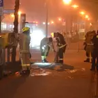 Mehrere Feuerwehrleute in Schutzkleidung arbeiten nachts auf einem Bürgersteig, einer hält einen Schlauch, ein anderer leuchtet mit einer Taschenlampe auf den Boden.