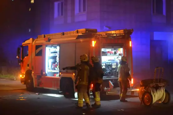 Drei Feuerwehrleute in Schutzkleidung stehen neben einem roten Feuerwehrauto mit geöffneten Fächern und eingeschalteten Lichtern bei Nacht.