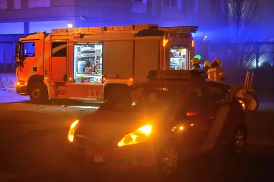 Rotes Feuerwehrfahrzeug mit geöffneten Fächern und mehreren Feuerwehrleuten in Schutzkleidung bei Nacht, davor ein Einsatzfahrzeug mit eingeschaltetem Warnlicht.
