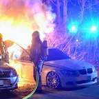 Zwei Feuerwehrleute mit Atemschutzgeräten löschen brennendes Auto, daneben ein weiteres geparktes Auto, Blaulicht im Hintergrund.