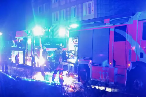 Feuerwehrfahrzeuge mit eingeschalteten Blaulichtern vor einem mehrstöckigen Gebäude bei Nacht, Feuerwehrleute in Schutzkleidung stehen davor.