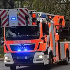 Rote Drehleiter mit eingeschaltetem Blaulicht auf einer Straße, umgeben von Bäumen.