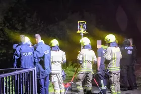 Mehrere Feuerwehrleute in Schutzkleidung und Polizisten stehen nachts an einem beleuchteten Ort im Freien, eine Person mit verpixeltem Gesicht ist dabei.