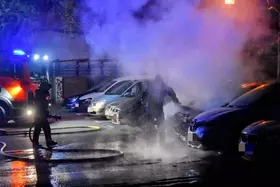Feuerwehrleute löschen nachts ein brennendes Auto auf einem Parkplatz, Rauch steigt auf, mehrere geparkte Autos sichtbar.