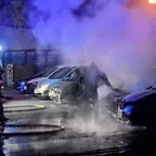 Feuerwehrleute löschen nachts ein brennendes Auto auf einem Parkplatz, Rauch steigt auf, mehrere geparkte Autos sichtbar.