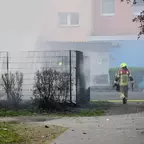 Feuerwehrmann in Schutzkleidung geht auf einen Brand an einem Zaun mit rauchenden Büschen zu.