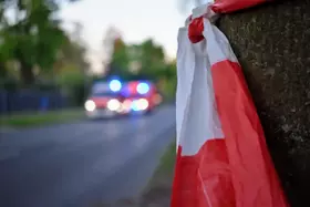 Rot-weißes Absperrband an einem Baumstamm am Straßenrand, im Hintergrund unscharf ein Fahrzeug mit Blaulicht.