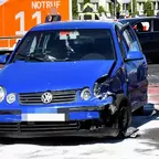 Blauer Volkswagen Kleinwagen mit stark beschädigtem rechtem Vorderteil, im Hintergrund Rettungswagen mit 'NOTRUF 112'.