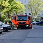 Feuerwehrfahrzeug mit geöffneter Beifahrertür steht auf einer Straße zwischen geparkten Autos und Bäumen vor einem Wohnhaus.