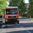 Orangerotes Feuerwehrfahrzeug der Marke MAN auf einer Straße, links beschädigtes weißes Auto mit abgerissenem Stoßfänger am Straßenrand.