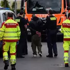 Mehrere Einsatzkräfte der Feuerwehr und Polizei begleiten eine Person mit gefesselten Händen auf einer Straße vor einem Feuerwehrfahrzeug.