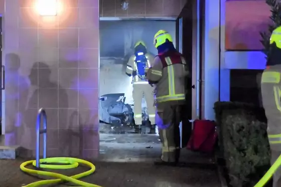 Zwei Feuerwehrleute in Schutzkleidung und Helmen betreten einen Raum mit Brandspuren, ein gelber Feuerwehrschlauch liegt auf dem Boden.