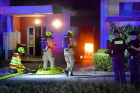 Drei Feuerwehrleute in Schutzkleidung und Atemschutz vor einem brennenden Müllraum, zwei Polizisten in dunkler Kleidung stehen daneben.