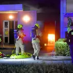 Drei Feuerwehrleute in Schutzkleidung und Atemschutz vor einem brennenden Müllraum, zwei Polizisten in dunkler Kleidung stehen daneben.