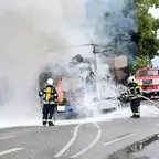Zwei Feuerwehrleute löschen einen brennenden LKW auf einer Straße, Rauch steigt auf, im Hintergrund ein rotes Feuerwehrauto und Häuser.