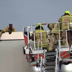 Drei Feuerwehrleute in Schutzkleidung und gelben Helmen auf einer Drehleiter neben einer Gebäudekante, ein Feuerwehrmann lehnt sich über die Dachkante und hält ein Blech fest
