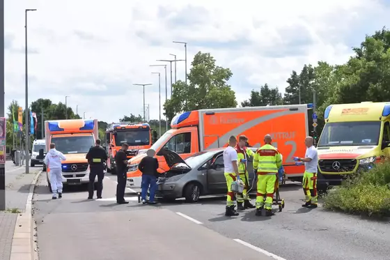 Mehrere Rettungskräfte in gelber und weißer Schutzkleidung stehen neben einem grauen Auto mit geöffneter Motorhaube auf einer Straße, im Hintergrund mehrere Rettungswagen.