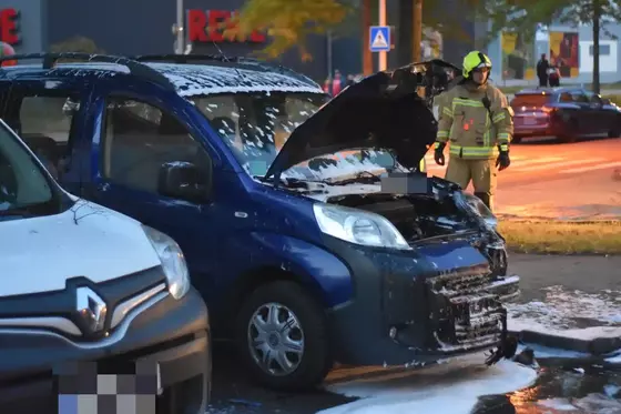 Blauer PKW mit geöffneter Motorhaube, bedeckt mit Löschschaum, daneben Feuerwehrmann in Schutzkleidung auf Straße bei Dämmerung.