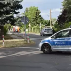 Polizeiauto mit blauem Streifen steht an einer Kreuzung in einer Wohnstraße mit Bäumen und Verkehrsschildern, im Hintergrund ein Fahrzeug der Feuerwehr.