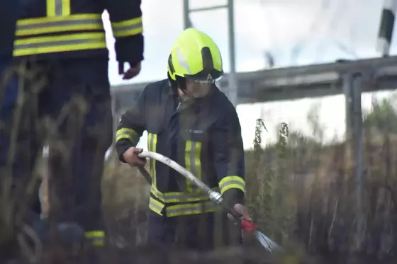 Feuerwehrmann mit gelbem Helm und Schutzkleidung hält Löschschlauch in einem abgebrannten Feld.