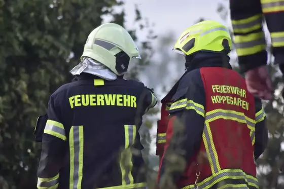 Zwei Feuerwehrleute in Schutzkleidung, einer mit gelbem Helm und rotem Einsatzanzug, der andere mit weißem Helm und schwarzem Anzug, von hinten aufgenommen.