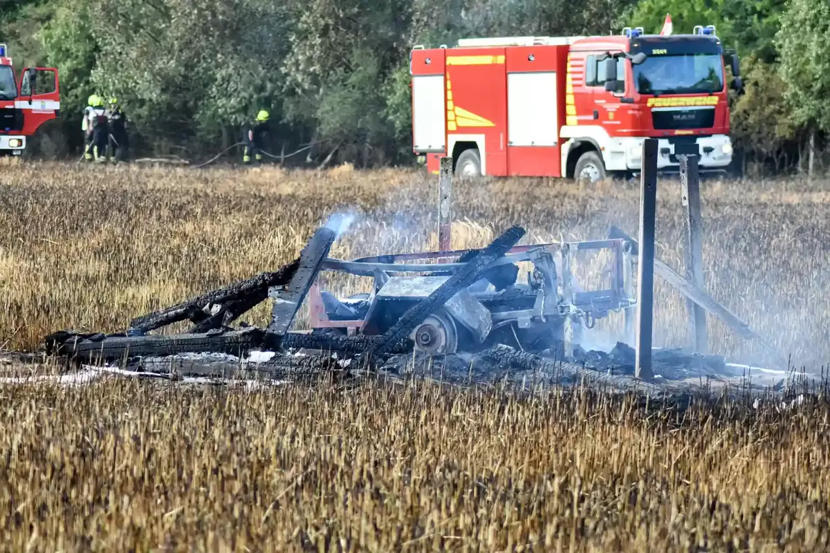 Abgebrannter Hochstand mit rauchenden, verkohlten Holzresten auf einem abgebrannten Feld, im Hintergrund zwei rote Feuerwehrfahrzeuge und Feuerwehrleute.